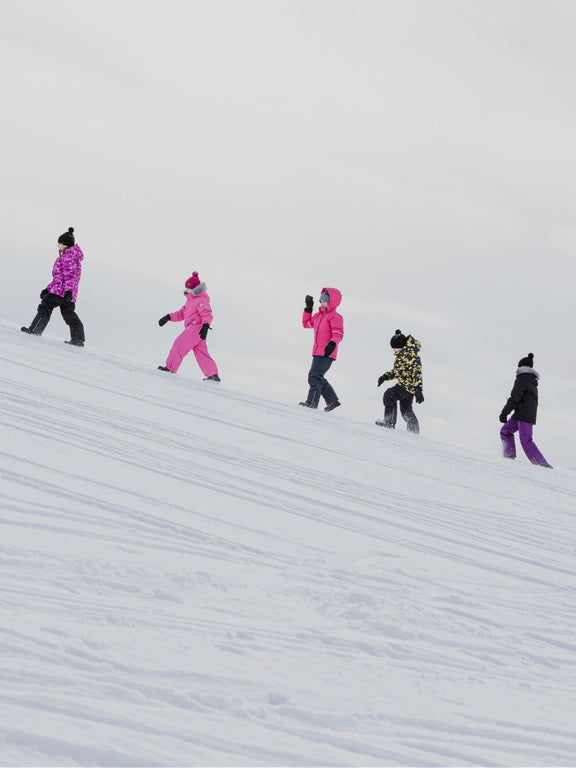 Viisi ihmistä värikkäissä talvivaatteissa kävelemässä lumisella rinteellä